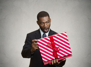 Closeup portrait happy excited surprised young businessman about to open unwrap red gift box isolated grey background, enjoying his present. Positive human emotions, facial expression feeling attitude